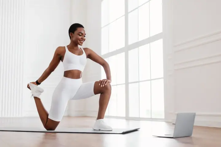 Femme en tenue de sport blanche effectuant un exercice quadriceps sur un tapis, guidée par un ordinateur portable.