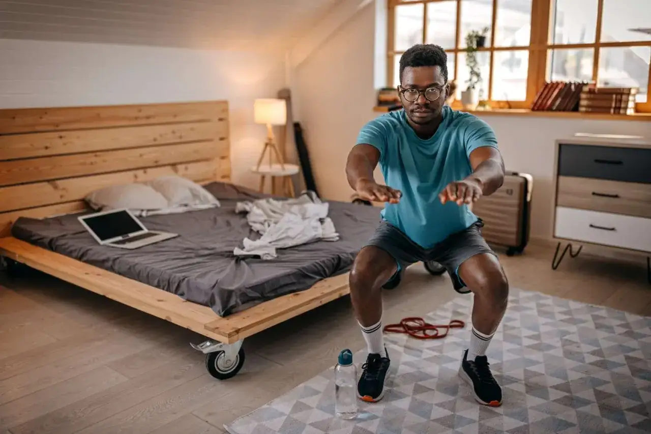 Un homme fait des squats pour son renforcement musculaire &agrave; la maison, pr&egrave;s de son lit.