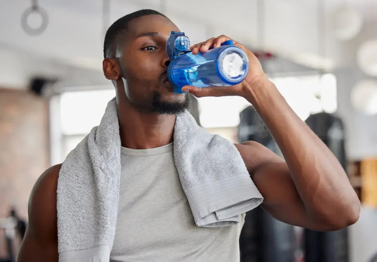 Un homme boit de l'eau d'une bouteille bleue après son entraînement. Il est important de savoir quelle quantité d'eau boire par jour pour rester hydraté.