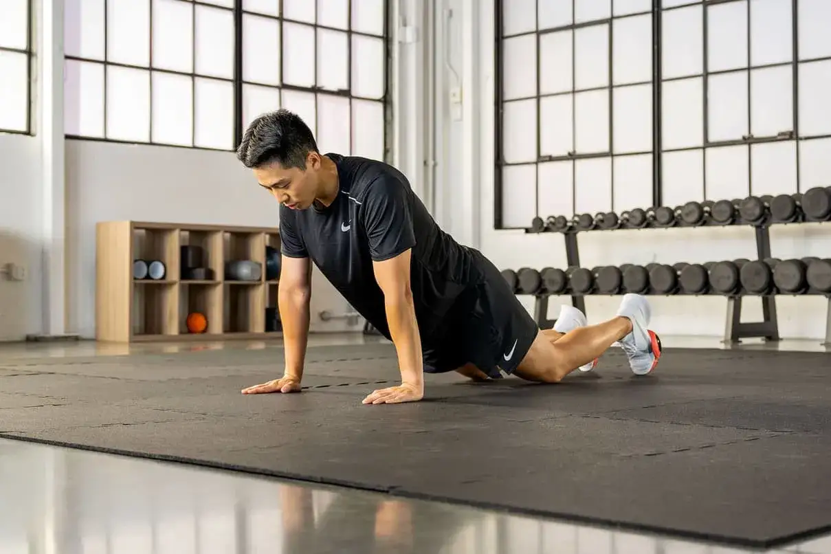 Un homme fait la planche sur un tapis de gym, les genoux au sol, dans une salle de sport avec des halt&egrave;res en arri&egrave;re-plan.