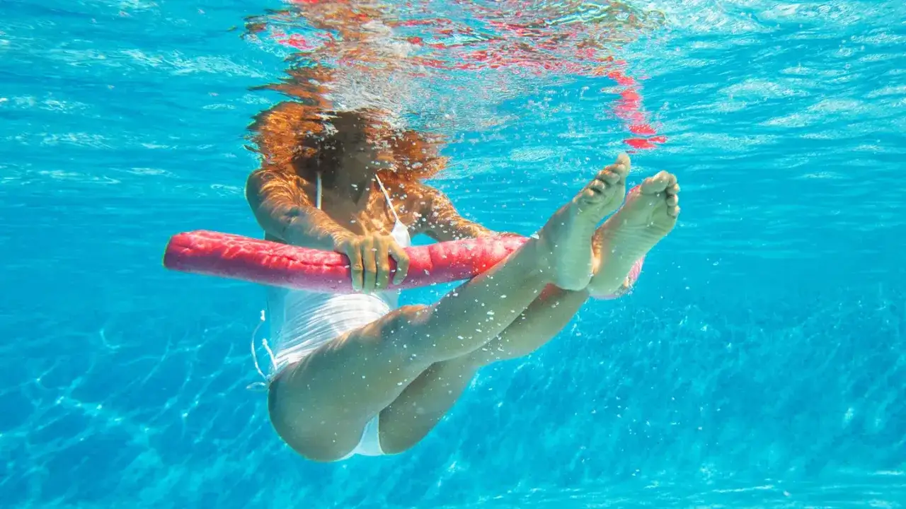 Femme sous l'eau tenant un flotteur rose pour un exercice aquagym. Ses jambes sont levées, ses pieds visibles.