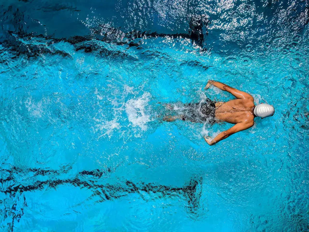 Un nageur en bonnet blanc nage en crawl dans une piscine bleue, créant des remous.