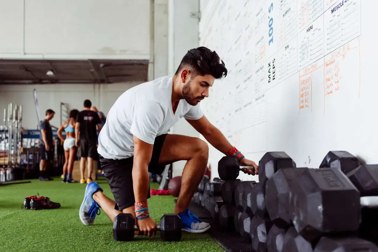 Un homme s'accroupit pour attraper des haltères, prêt pour ses exercices musculation. D'autres personnes s'entraînent en arrière-plan.