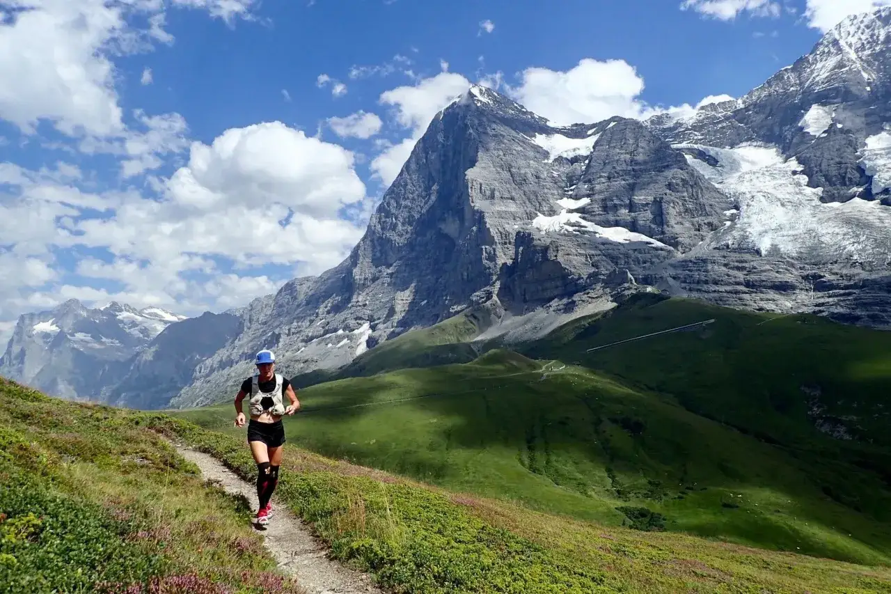 Une femme en plein effort de rando course sur un sentier de montagne, avec des sommets enneig&eacute;s en toile de fond.